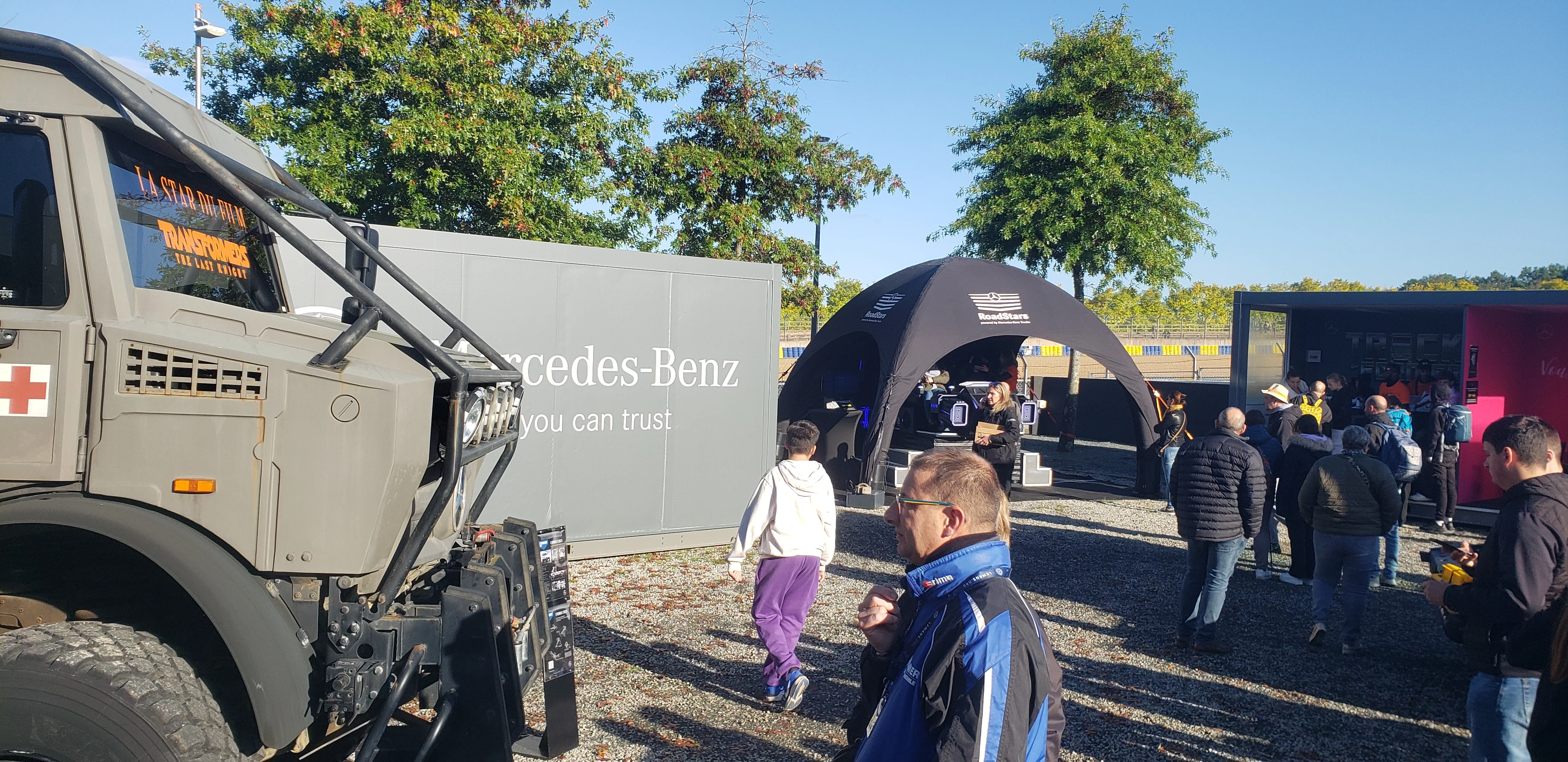 Paddock avec Unimog militaire stand Mercedes et tente RoadStars du simulateur navette VR au fond