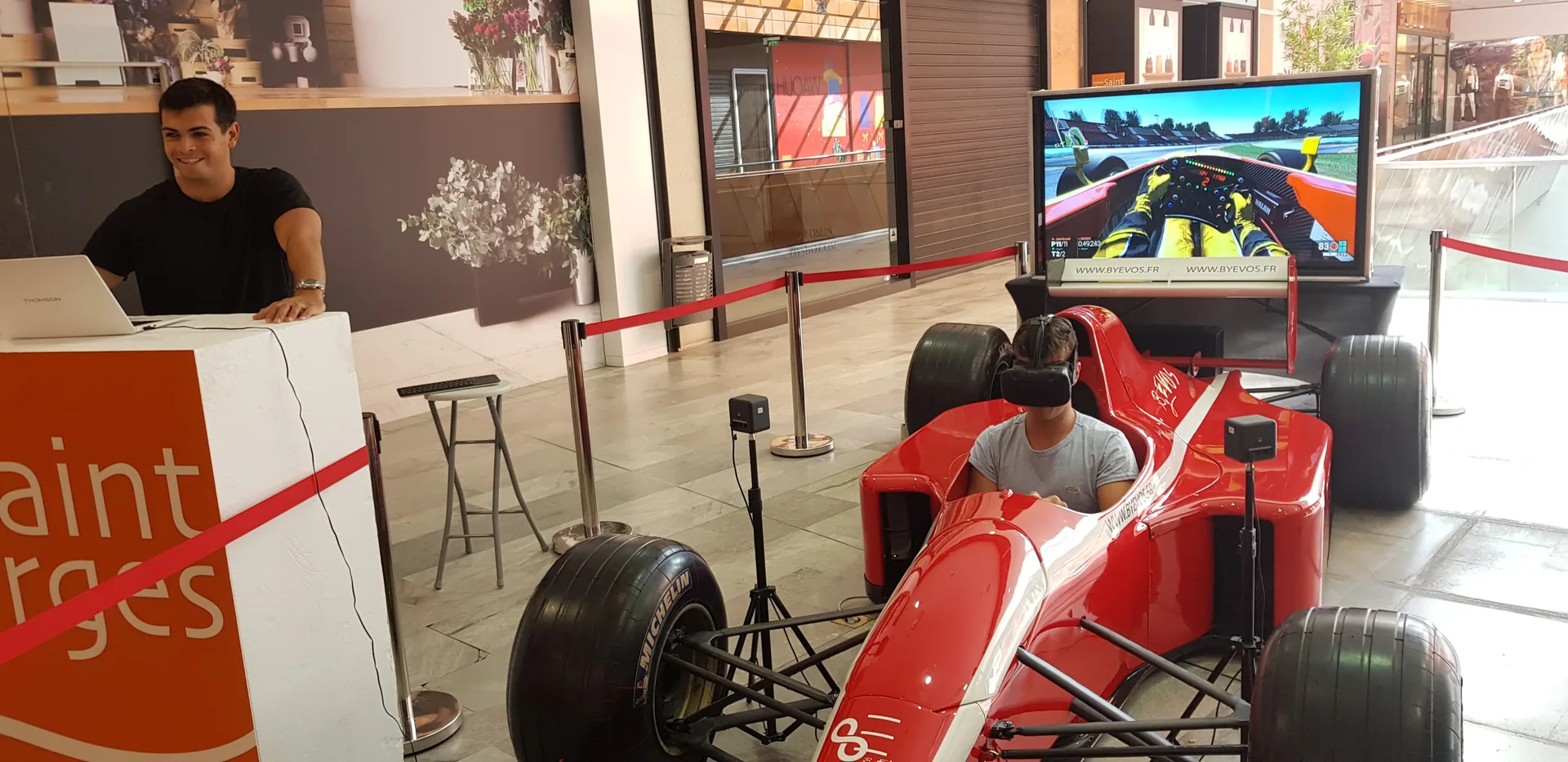 Animateur souriant à la borne d'accueil avec participant casqué VR dans le cockpit F1 à l'Espace Saint-Georges