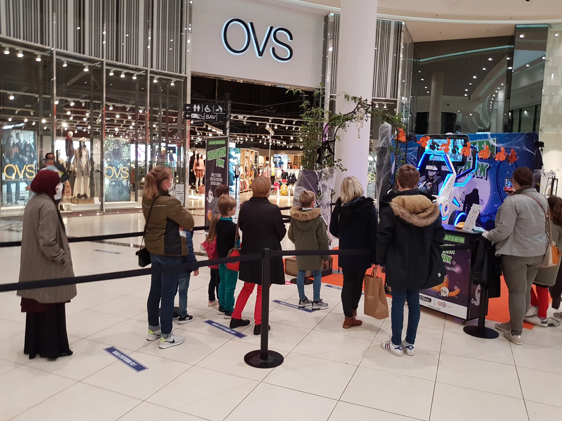Famille avec enfants en file d'attente devant le simulateur SOL 360 espacée par les marquages distanciation au sol face à OVS
