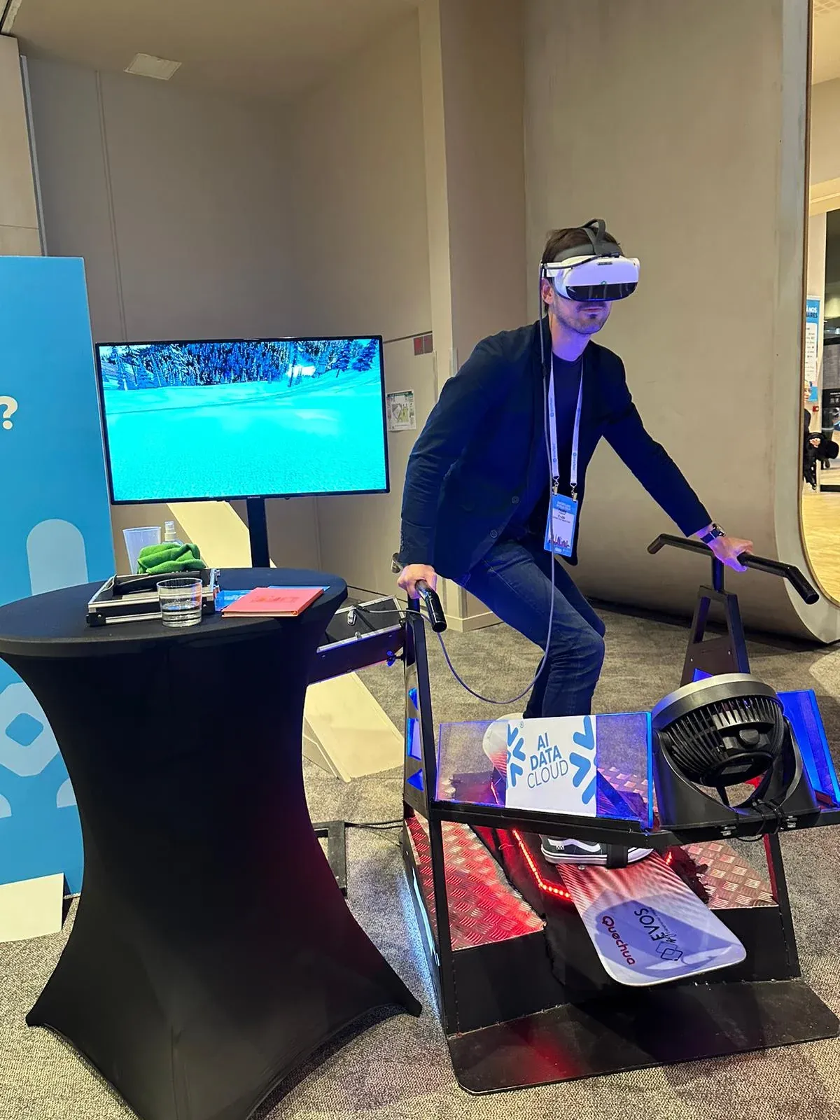 Homme en blazer noir et casque VR debout sur un simulateur snowboard avec écran affichant un paysage enneigé sur un salon professionnel