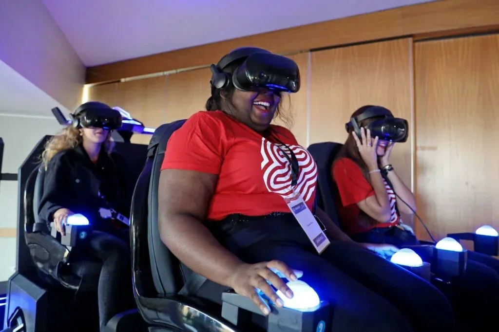 Trois participantes en casque VR à bord de la navette Evos dont la passagère centrale sourit largement avec badges visiteur dans une salle aux murs boisés et éclairage violet