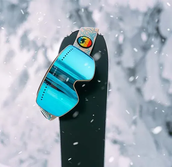 Masque de ski à verres bleus miroir posé sur un snowboard noir avec flocons de neige en arrière-plan