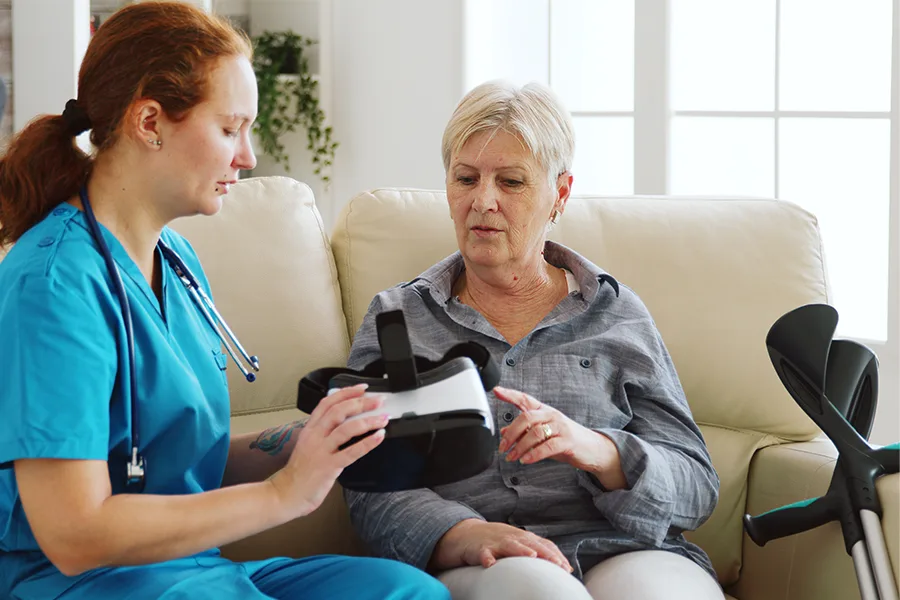 Aide-soignante présentant un casque VR à une résidente assise sur un canapé